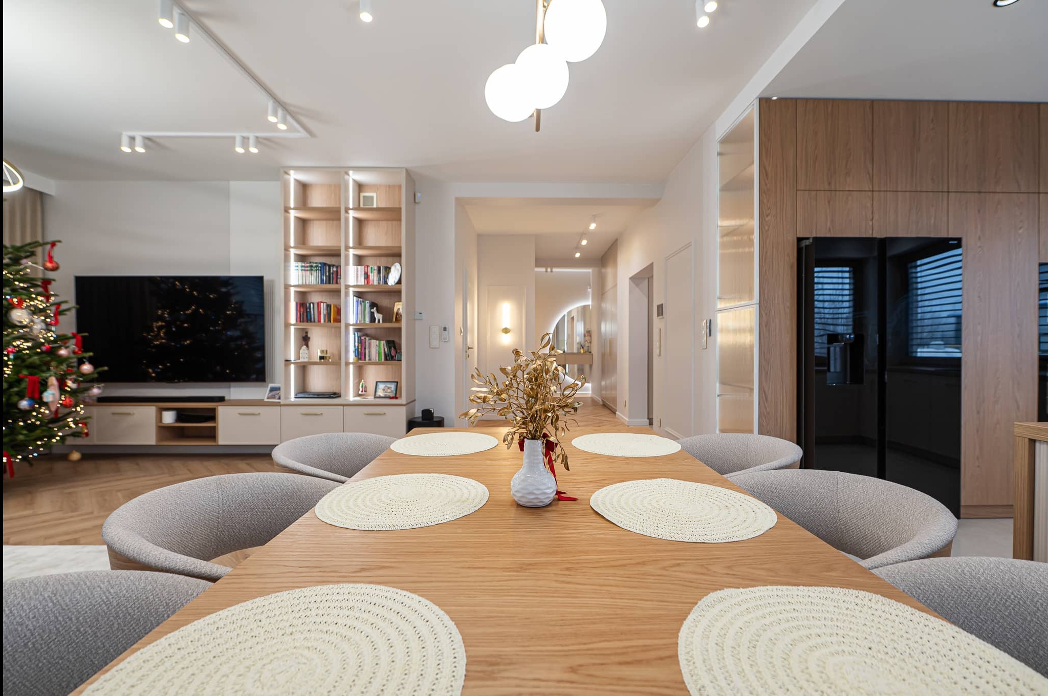 Jasne wnętrze jadalni z drewnianym stołem, okrągłymi podkładkami i szarymi krzesłami. W tle regał z książkami, telewizor i choinka. Nowoczesne oświetlenie.