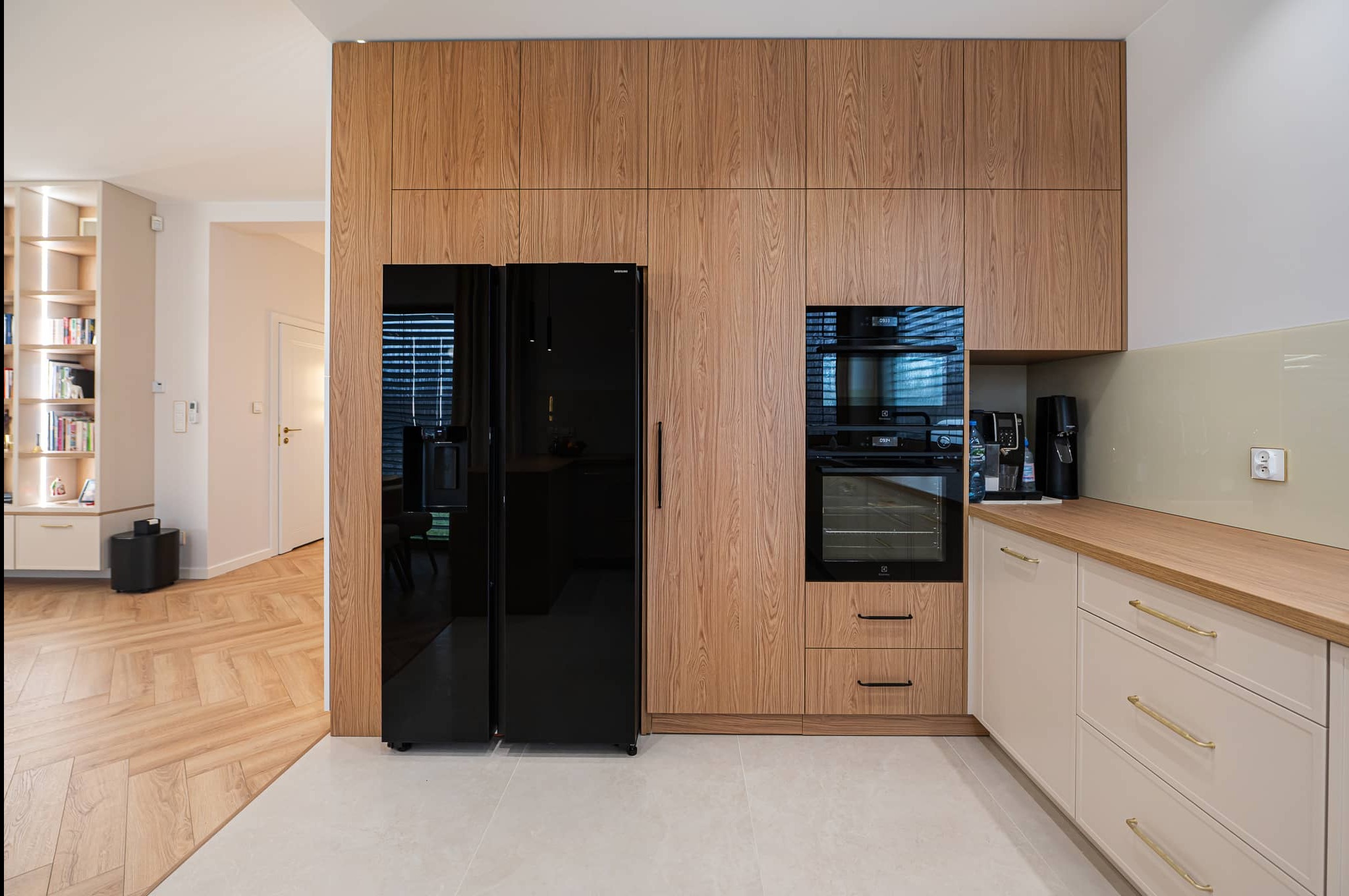Nowoczesna kuchnia z drewnianymi szafkami, czarną lodówką side-by-side i piekarnikiem w zabudowie. Jasne blaty i podłoga. Widok na salon z parkietem w jodełkę.