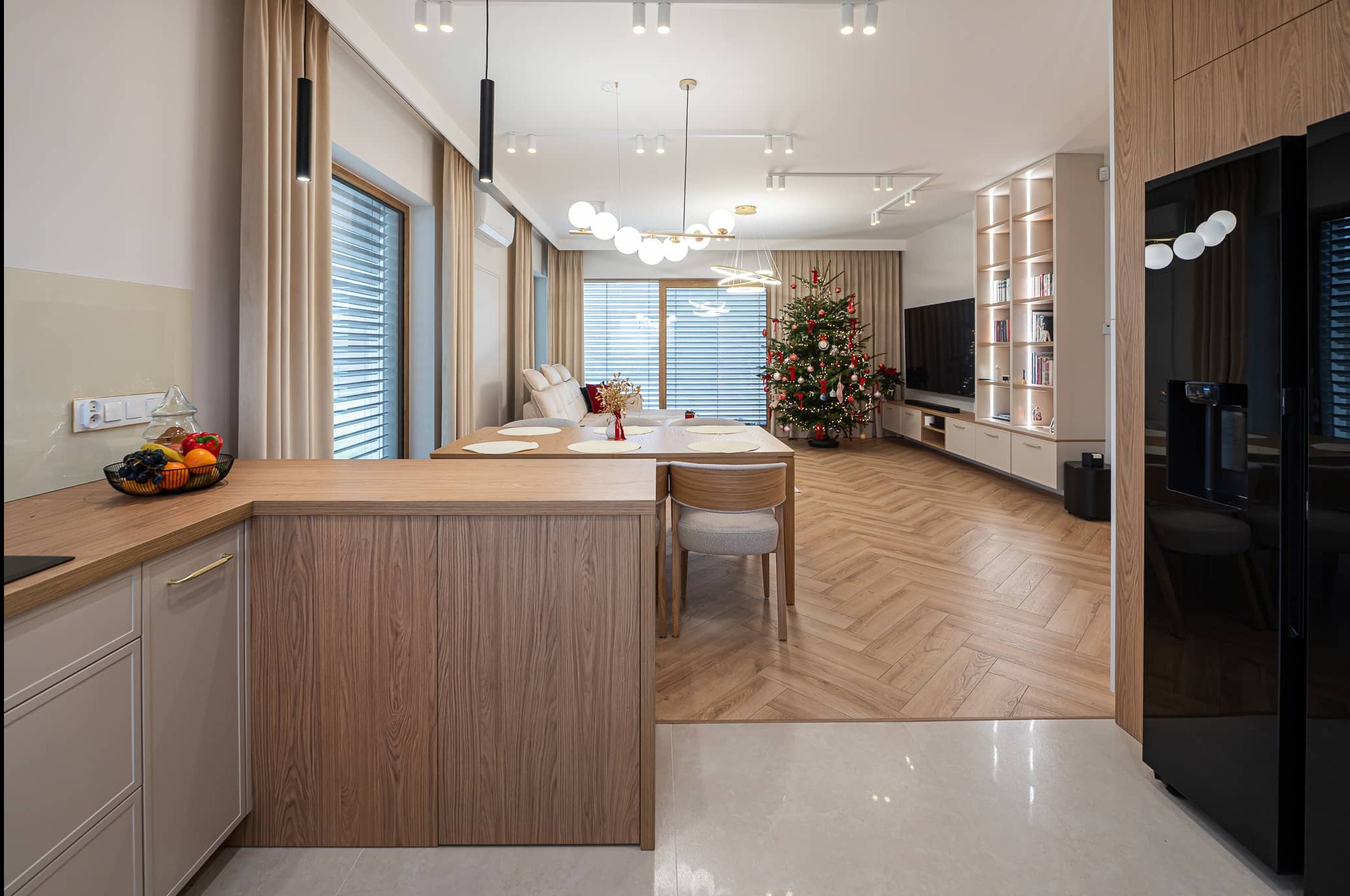 Nowoczesne wnętrze z drewnianą podłogą w jodełkę, wyspą kuchenną i świąteczną choinką w tle. Oświetlenie punktowe i wiszące lampy dodają elegancji.