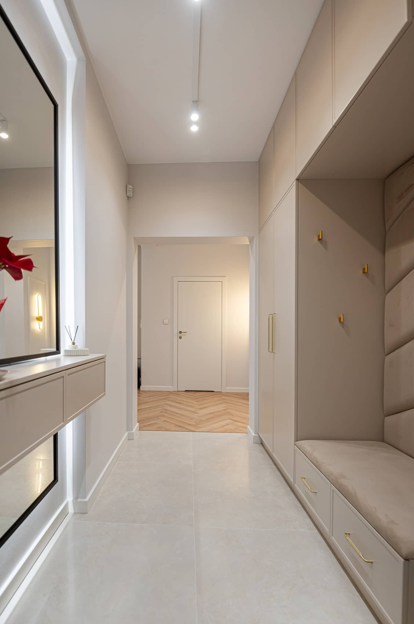 Nowoczesny przedpokój w jasnych barwach: lustro z podświetleniem, siedzisko z wieszakami, szafa w zabudowie, płytki podłogowe i parkiet w jodełkę w tle.