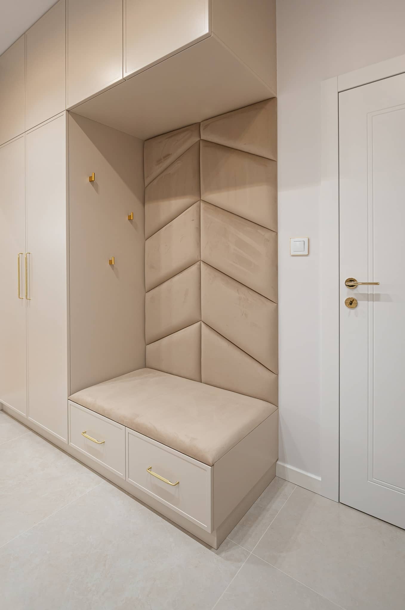 Beżowa zabudowa przedpokoju z siedziskiem i panelami ściennymi w geometryczny wzór. Złote uchwyty i haczyki dodają elegancji. Minimalistyczny design.
