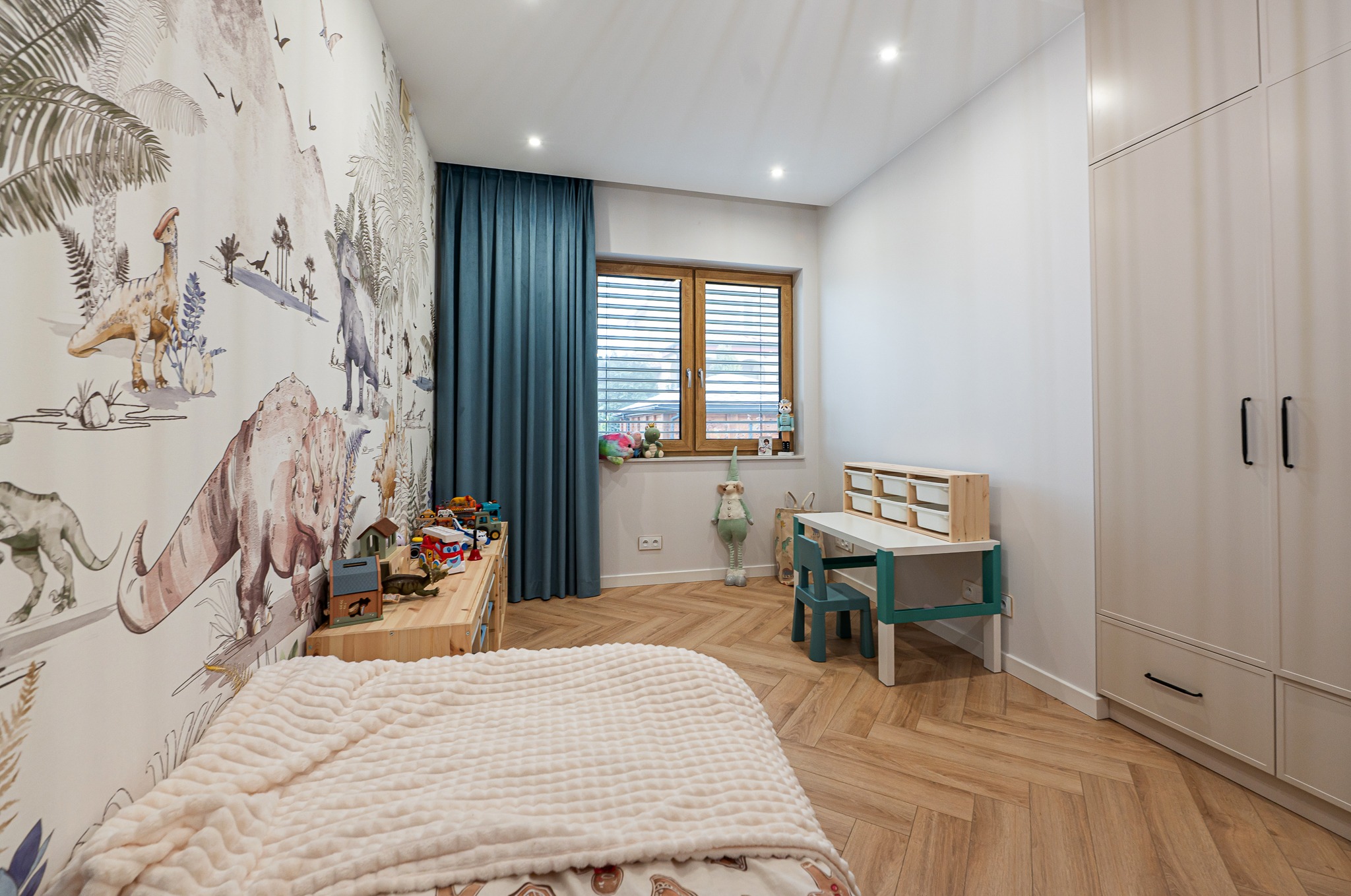 Pokój dziecięcy z tapetą w dinozaury, drewnianą podłogą w jodełkę i biurkiem. Uwagę przykuwa pluszowy koc na łóżku i zabawki na komodzie.