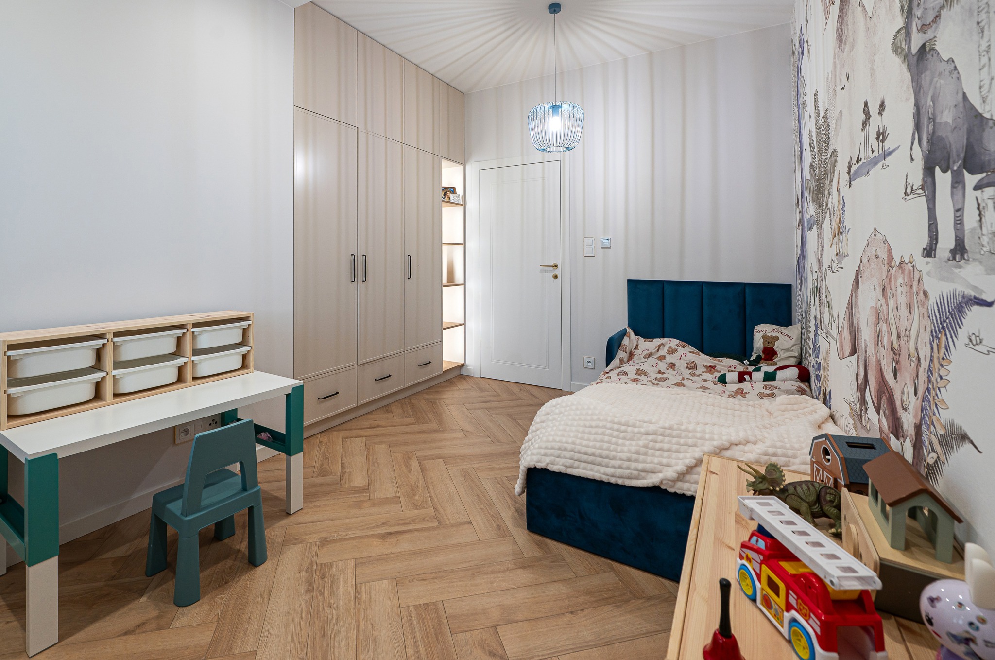 Pokój dziecięcy z łóżkiem z niebieskim zagłówkiem, biurkiem z pojemnikami, szafą w zabudowie i tapetą w dinozaury. Parkiet ułożony w jodełkę.