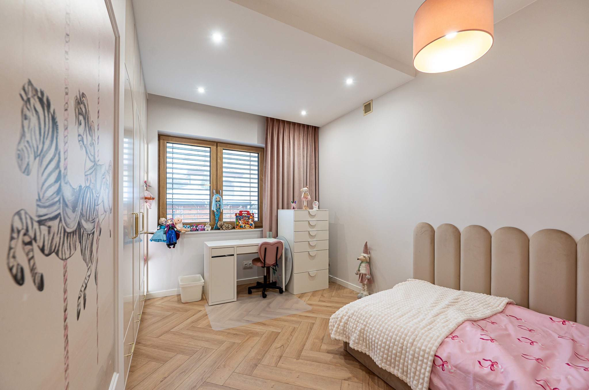 Pokój dziecięcy w Cekanowie: łóżko z beżowym zagłówkiem, biurko przy oknie, szafa z motywem karuzeli, parkiet w jodełkę. Przytulne i funkcjonalne wnętrze.