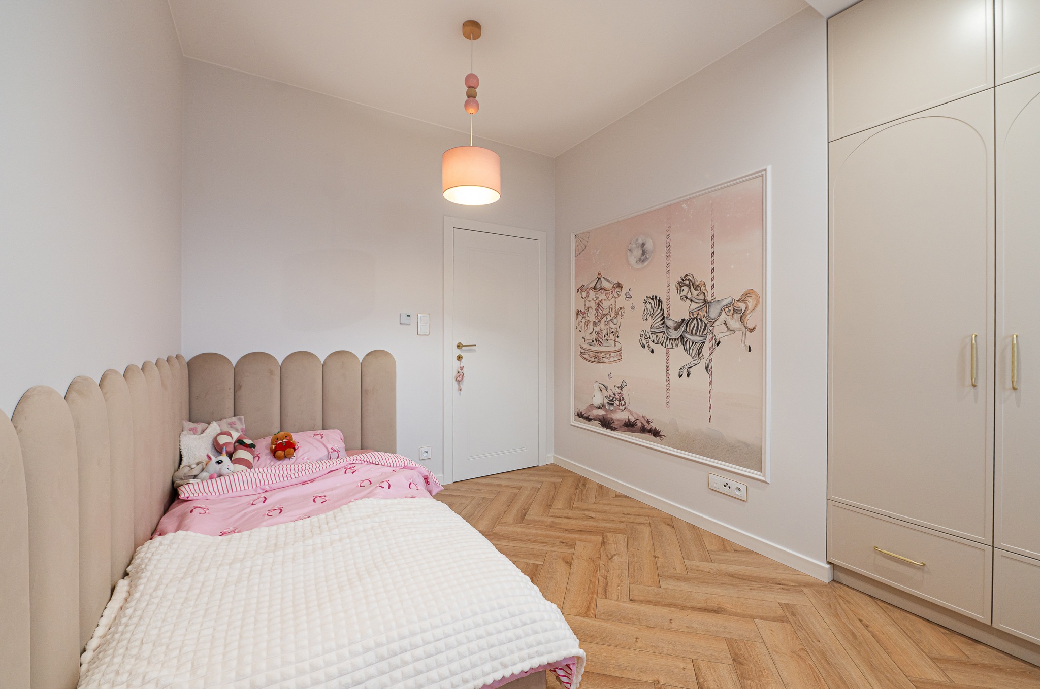 Pokój dziecięcy w pastelowych barwach: łóżko z pluszakami, tapeta z motywem karuzeli, parkiet w jodełkę i szafa z zaokrąglonymi frontami.