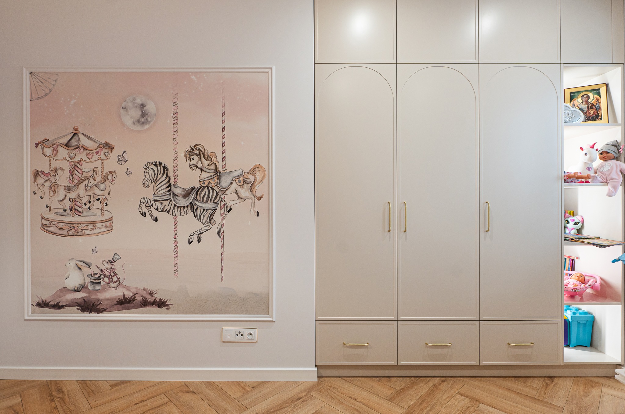 Pokój dziecięcy: pastelowa tapeta z karuzelą, duża szafa z zaokrąglonymi drzwiami i otwartą półką z zabawkami. Parkiet w jodełkę.
