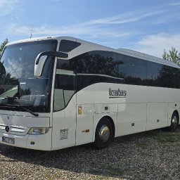 LEMBUS - Wynajem busów i autokarów, przewóz towarów oraz transport niskopodwoziowy - Biały autokar marki Mercedes z logo 'Lembus' zaparkowany na żwirowym parkingu, obok widoczne inne autobusy i zieleń drzew.