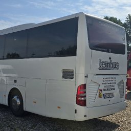 LEMBUS - Wynajem busów i autokarów, przewóz towarów oraz transport niskopodwoziowy - Tył białego autokaru Mercedes-Benz Tourismo firmy Lembus z widocznymi danymi kontaktowymi i tablicą rejestracyjną na tle żwirowego parkingu i fragmentu innego autobusu oraz czerwonej ciężarówki.
