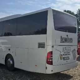 Tył białego autokaru Mercedes-Benz Tourismo firmy Lembus z widocznymi danymi kontaktowymi i tablicą rejestracyjną na tle żwirowego parkingu i fragmentu innego autobusu oraz czerwonej ciężar&oacute;wki.