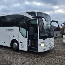 LEMBUS - Wynajem busów i autokarów, przewóz towarów oraz transport niskopodwoziowy - Biały autokar marki Mercedes-Benz z otwartymi drzwiami, widoczne logo Lembus, na parkingu z szarym żwirem, w tle inne autokary i budynek.