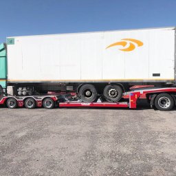 LEMBUS - Wynajem busów i autokarów, przewóz towarów oraz transport niskopodwoziowy - Ciężarówka z naczepą transportowana na niskopodwoziowej lawecie z zabezpieczonymi kołami, widok z boku na placu manewrowym w słoneczny dzień.