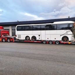 LEMBUS - Wynajem busów i autokarów, przewóz towarów oraz transport niskopodwoziowy - Czerwony ciągnik siodłowy z naczepą niskopodwoziową przewozi biały autokar z zabezpieczonym tyłem, na tle szarego, pochmurnego nieba i budynku z bramami garażowymi.