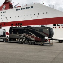 LEMBUS - Wynajem busów i autokarów, przewóz towarów oraz transport niskopodwoziowy - Ciężarówka transportuje dużą przyczepę kempingową na platformie, na tle ogromnego białego promu z czerwonym pasem i włoską flagą.