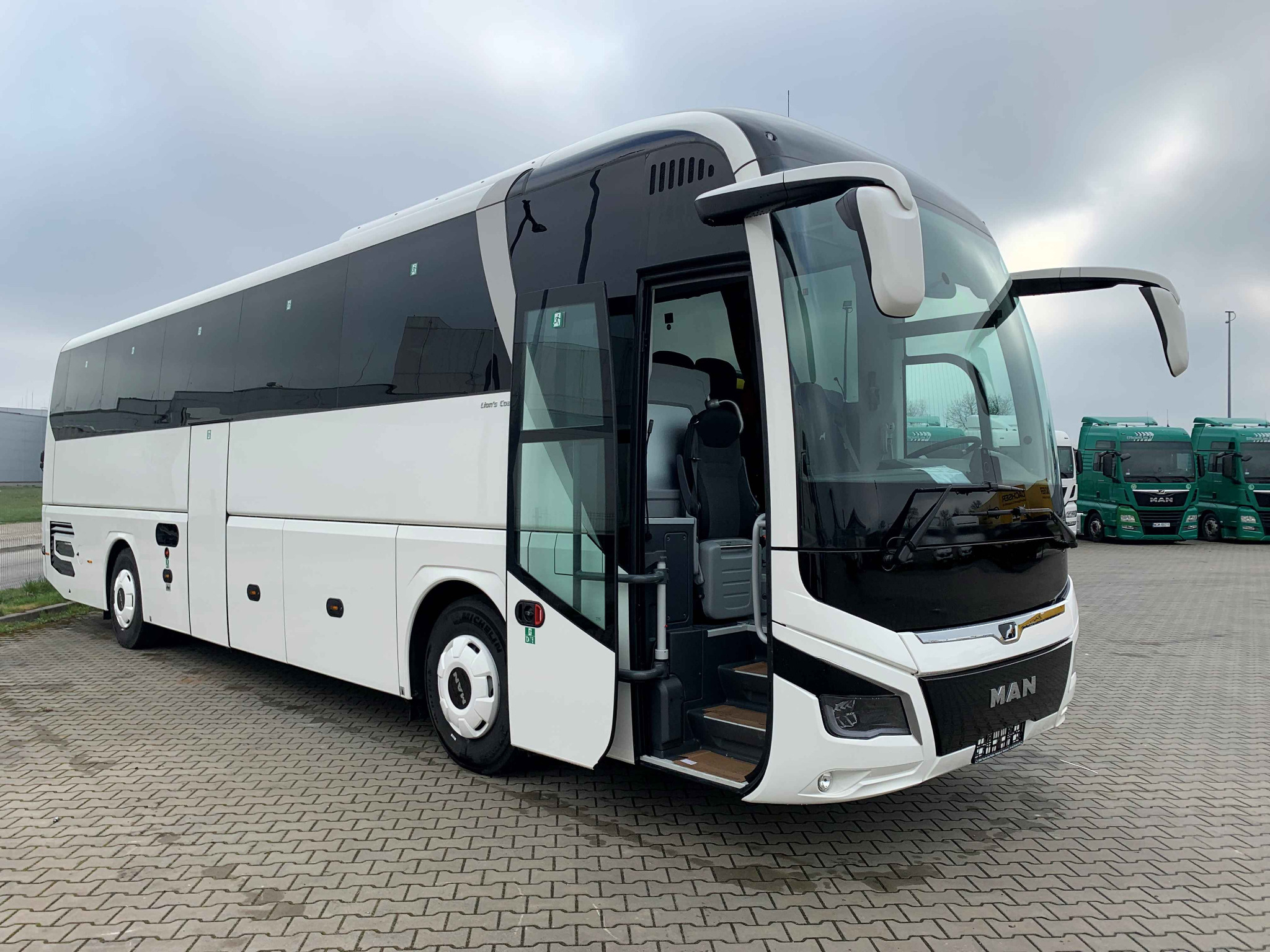 Nowoczesny, biało-czarny autokar MAN z otwartymi drzwiami wejściowymi, zaparkowany na brukowanej powierzchni, z rzędem zielonych ciężarówek MAN w tle pod pochmurnym niebem.