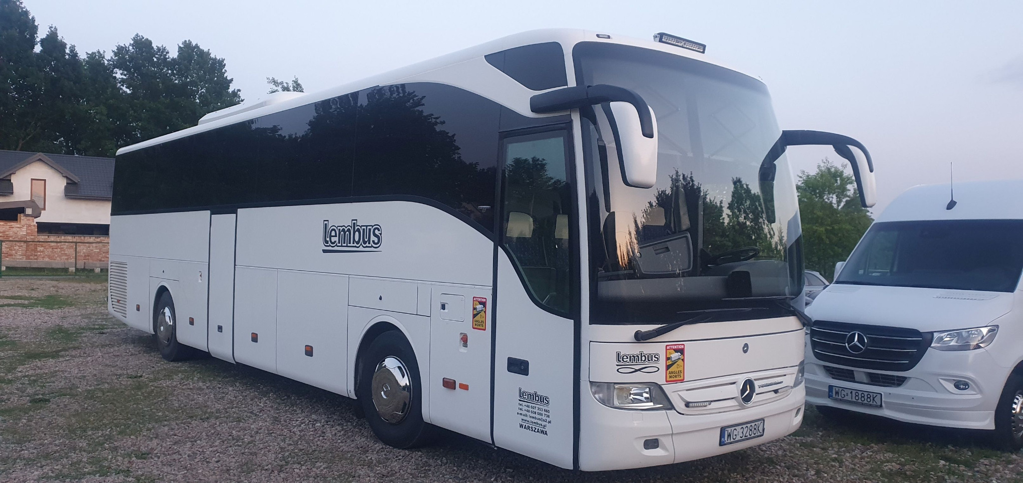 Biały autokar marki Mercedes-Benz z logo 'Lembus' na boku, zaparkowany na żwirowym terenie obok białego busa. Widoczne tablice rejestracyjne i oznaczenia ostrzegawcze.