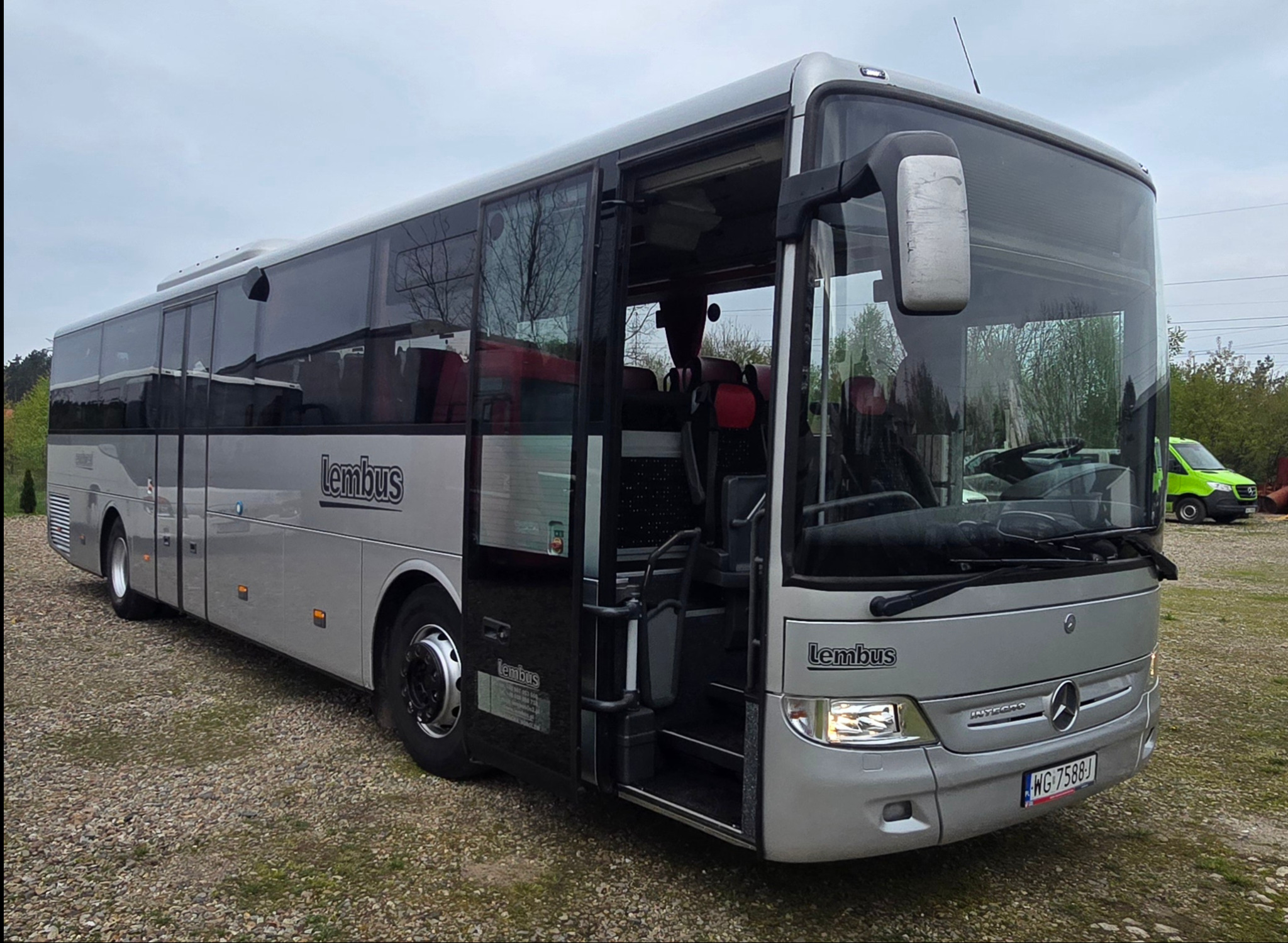 Srebrny autokar marki Mercedes z otwartymi drzwiami, widoczne siedzenia pasażerskie i logo 'Lembus' na boku, zaparkowany na żwirowym placu.