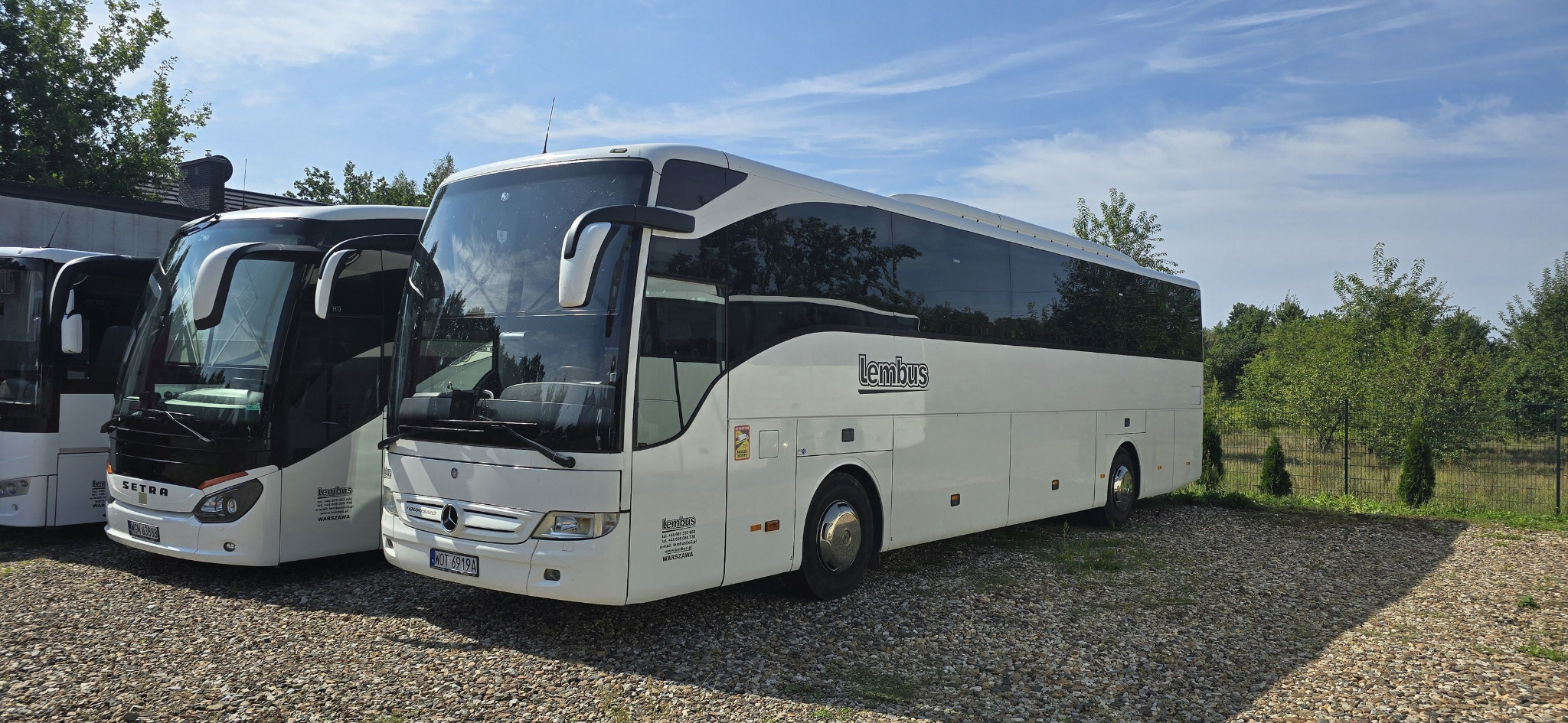Biały autokar marki Mercedes z logo 'Lembus' zaparkowany na żwirowym parkingu, obok widoczne inne autobusy i zieleń drzew.