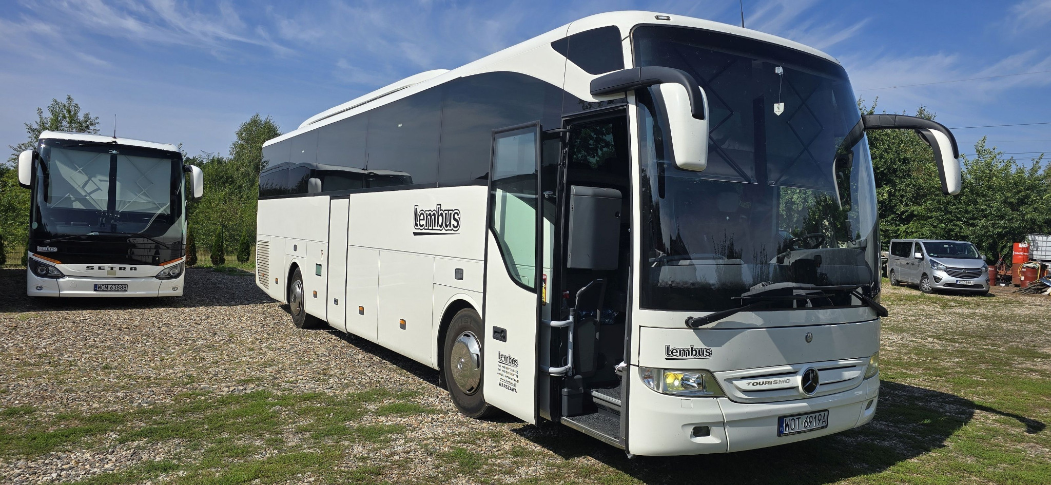 Biały autobus turystyczny z otwartymi drzwiami na żwirowym parkingu, z innym autobusem w tle i widocznym logo 'Lembus'.