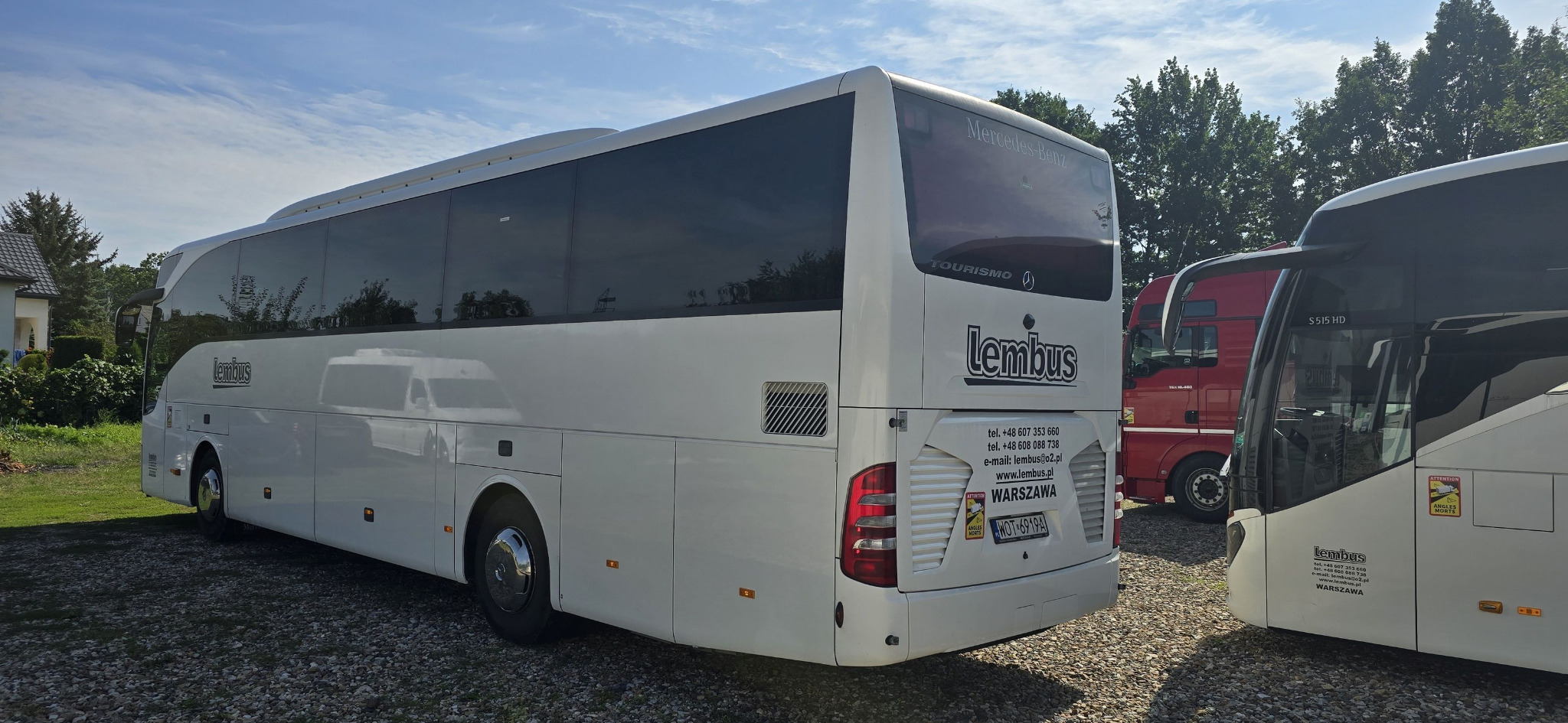 Tył białego autokaru Mercedes-Benz Tourismo firmy Lembus z widocznymi danymi kontaktowymi i tablicą rejestracyjną na tle żwirowego parkingu i fragmentu innego autobusu oraz czerwonej ciężarówki.