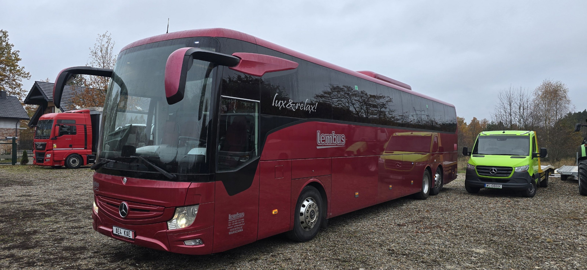 Czerwony autokar marki Mercedes-Benz firmy Lembus z napisem 'lux & relax!' na boku, zaparkowany na żwirowym placu obok czerwonej ciężarówki MAN i zielonej lawety Mercedes-Benz.