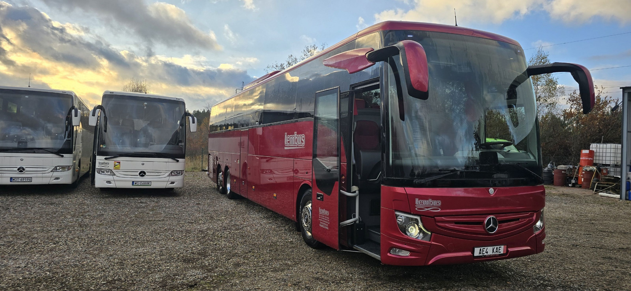 Czerwony autokar z otwartymi drzwiami na żwirowym parkingu, obok stoją dwa białe autokary, widoczne zbiorniki i beczki w tle, pochmurne niebo.