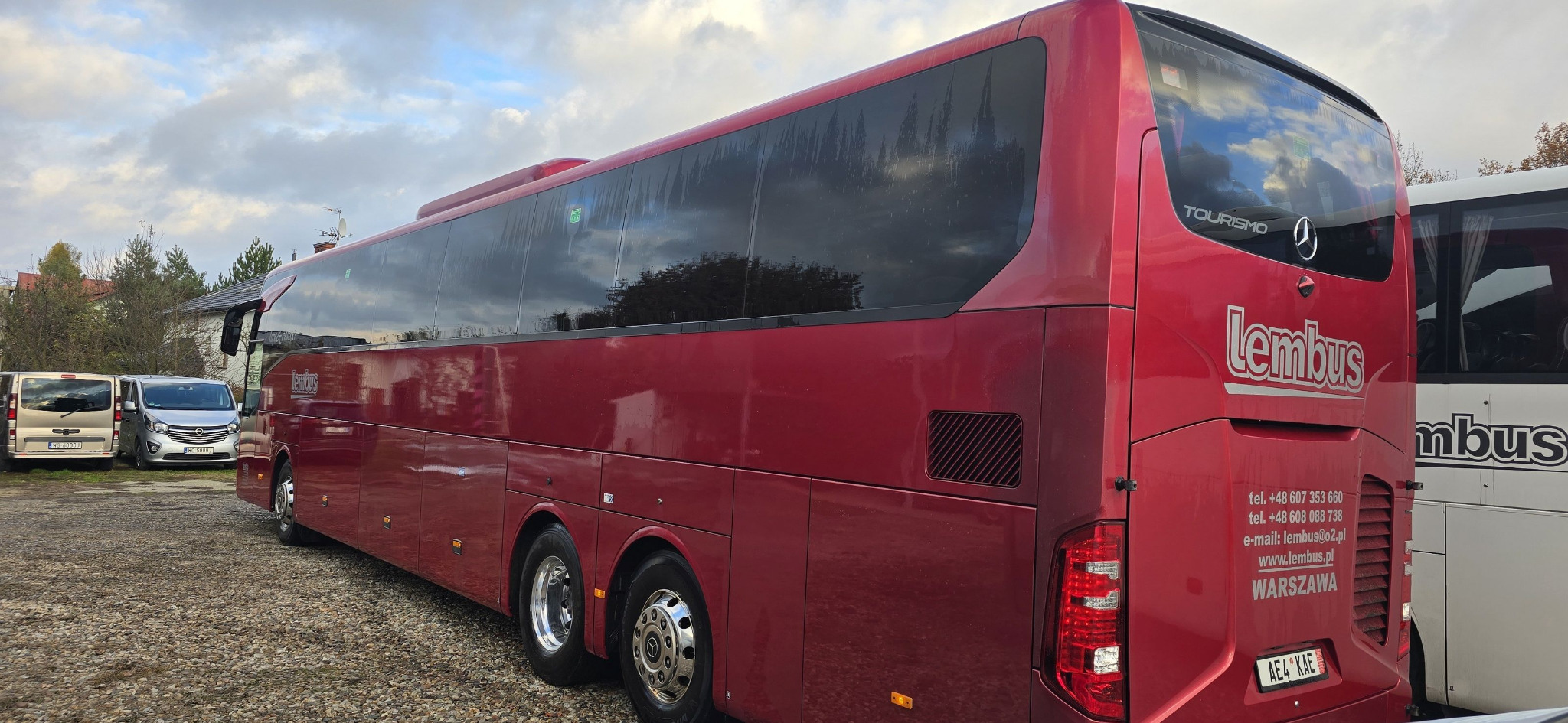 Czerwony autokar marki Lembus zaparkowany na żwirowym parkingu, widok z tyłu i boku, z widocznymi numerami telefonu i adresem e-mail firmy z Warszawy.