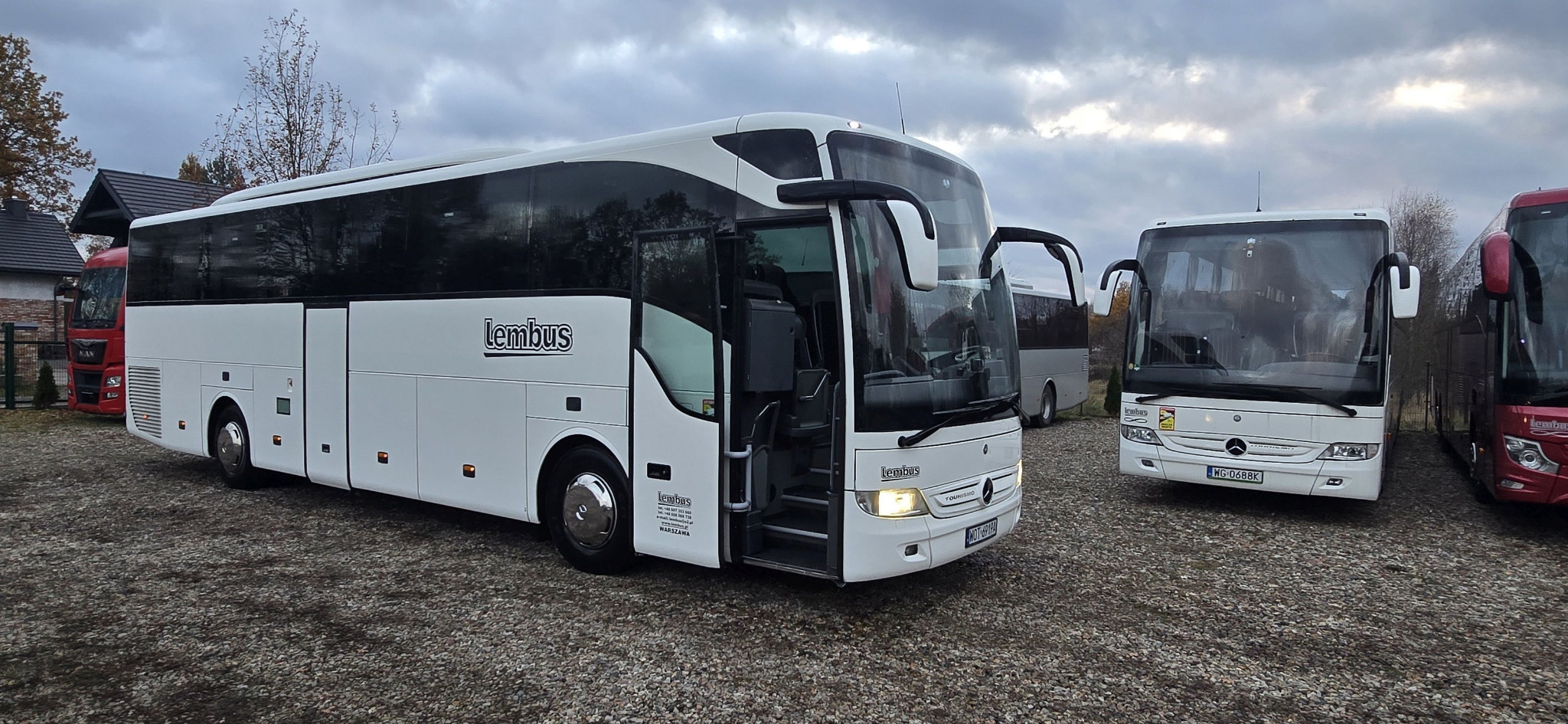 Biały autokar marki Mercedes-Benz z otwartymi drzwiami, widoczne logo Lembus, na parkingu z szarym żwirem, w tle inne autokary i budynek.