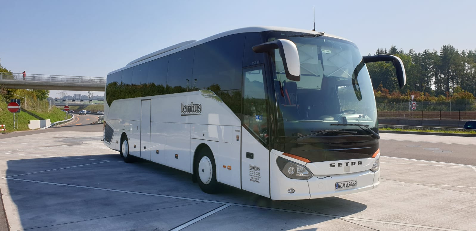 Biały autokar marki Setra z logo 'lembuss' zaparkowany na betonowym placu w słoneczny dzień, w tle wiadukt i zieleń.