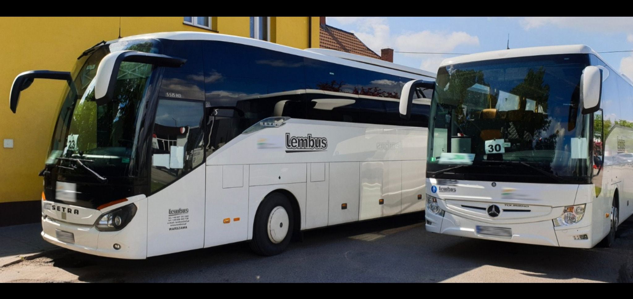 Dwa białe autokary marki Setra i Mercedes-Benz z logo 'Lembus' zaparkowane obok siebie na tle żółtego budynku i błękitnego nieba.