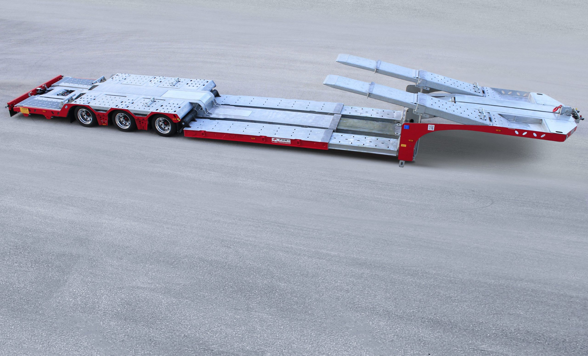 Nowa, pusta, niskopodwoziowa przyczepa lawetowa z rozkładanymi najazdami, pomalowana na czerwono i srebrno, stojąca na betonowym placu.