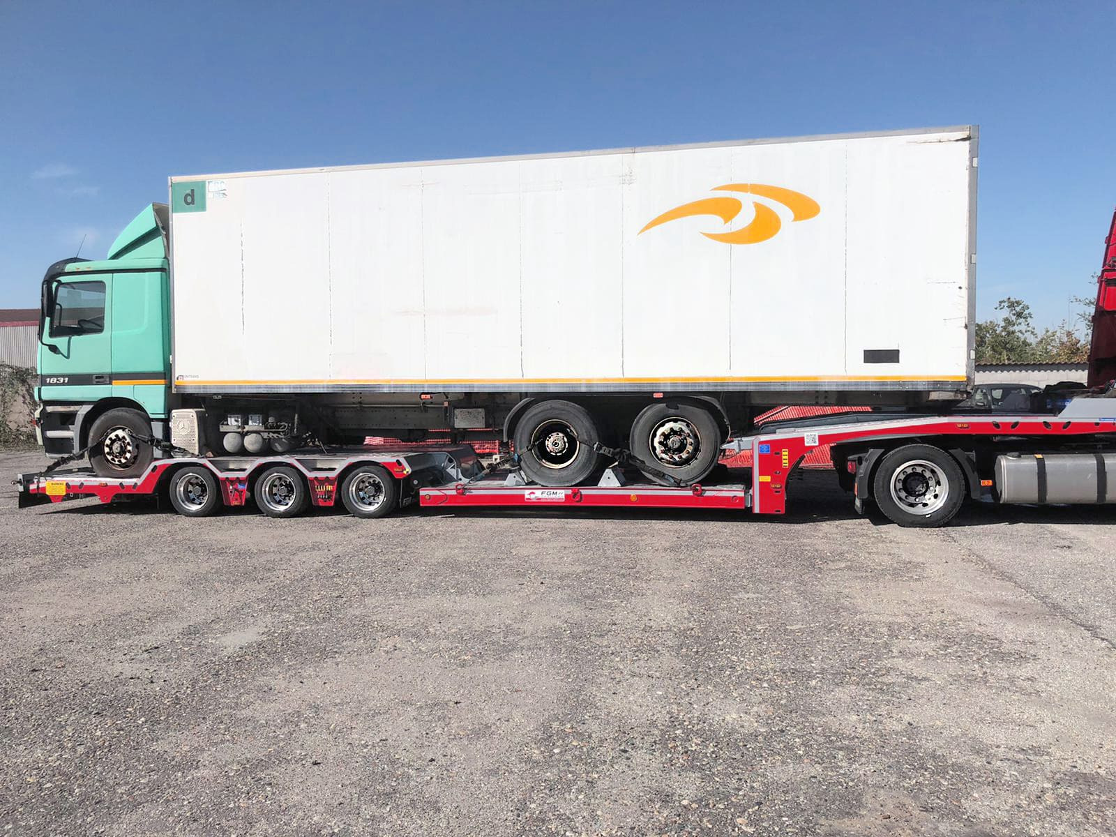 Ciężarówka z naczepą transportowana na niskopodwoziowej lawecie z zabezpieczonymi kołami, widok z boku na placu manewrowym w słoneczny dzień.