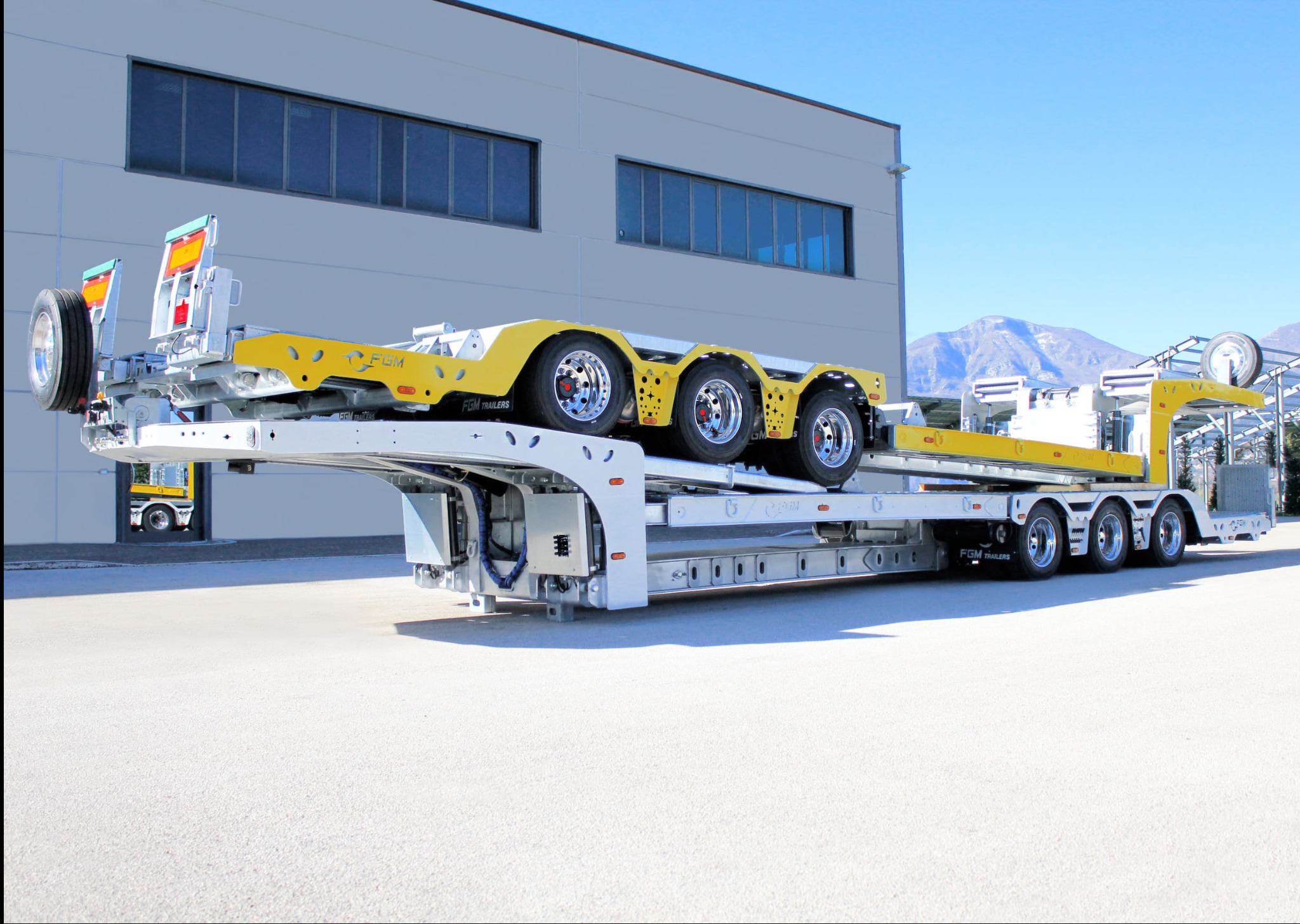 Nowoczesna, biało-żółta naczepa niskopodwoziowa FGM Trailers na betonowym placu, z budynkiem przemysłowym i górami w tle.