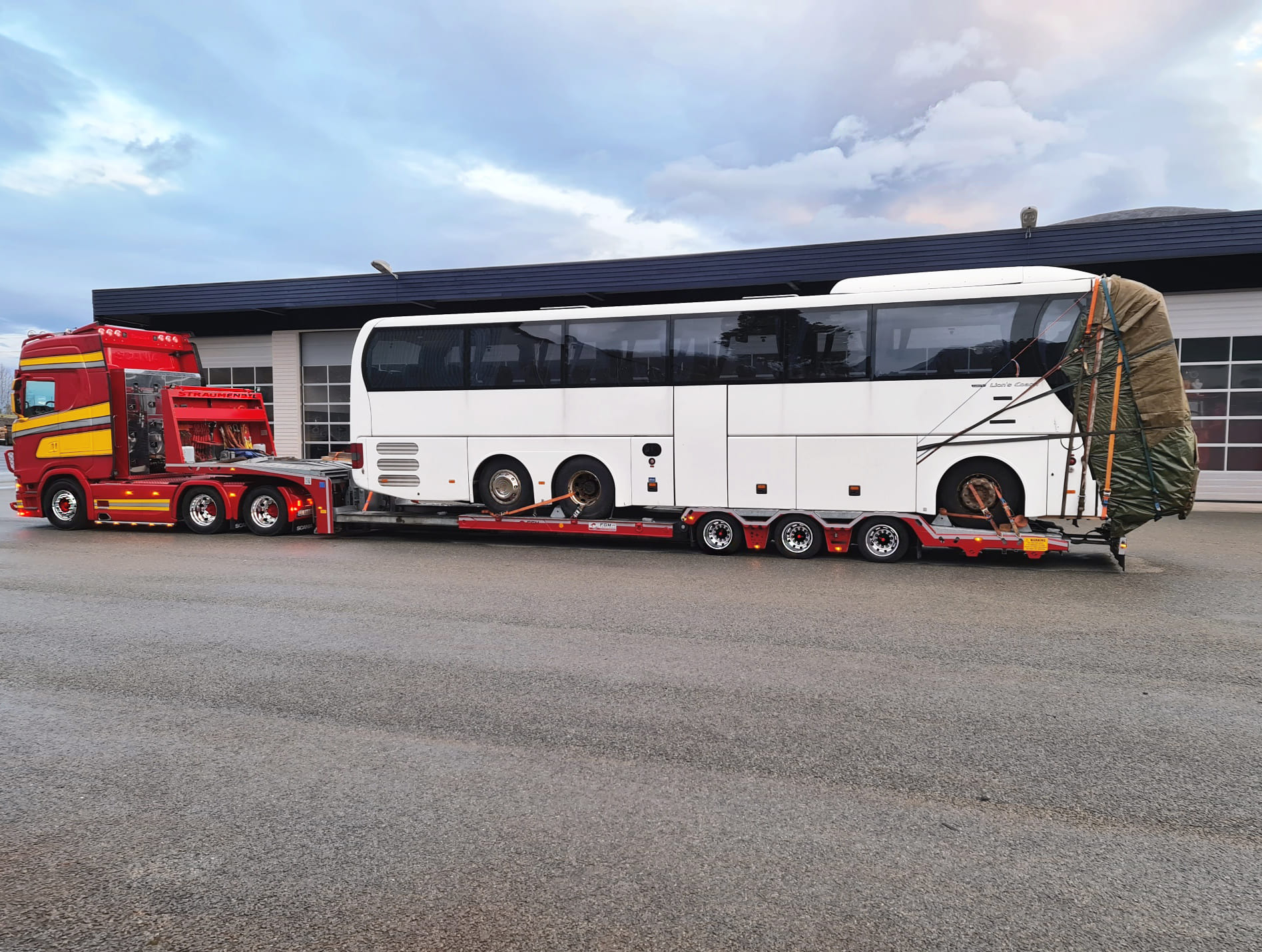 Czerwony ciągnik siodłowy z naczepą niskopodwoziową przewozi biały autokar z zabezpieczonym tyłem, na tle szarego, pochmurnego nieba i budynku z bramami garażowymi.