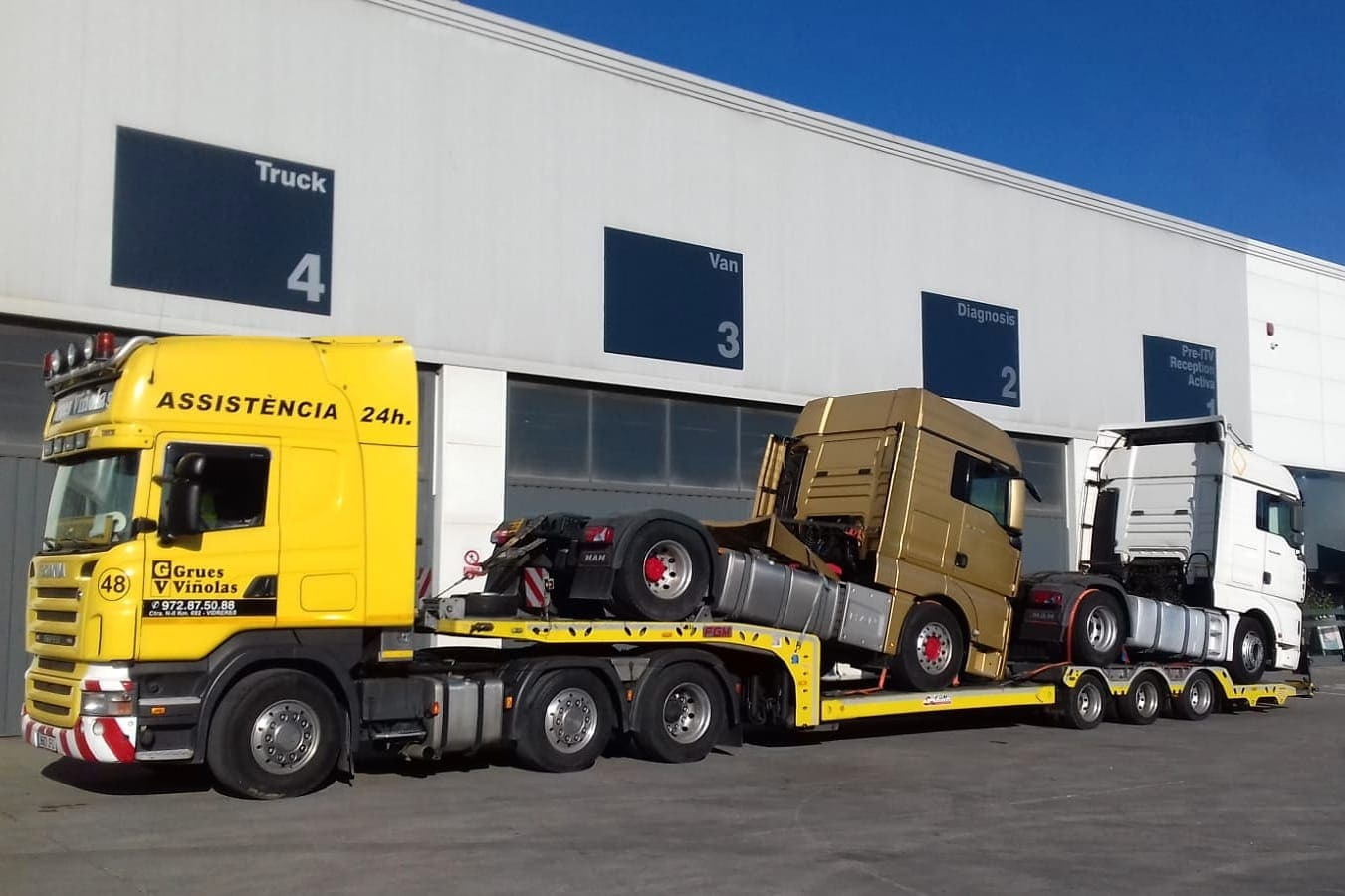 Żółta ciężarówka z napisem 'ASSISTÈNCIA 24h.' przewozi dwa ciągniki siodłowe, jeden złoty, drugi biały, na niskopodwoziowej platformie przed budynkiem z oznaczeniami 'Truck 4', 'Van 3', 'Diagnosis...