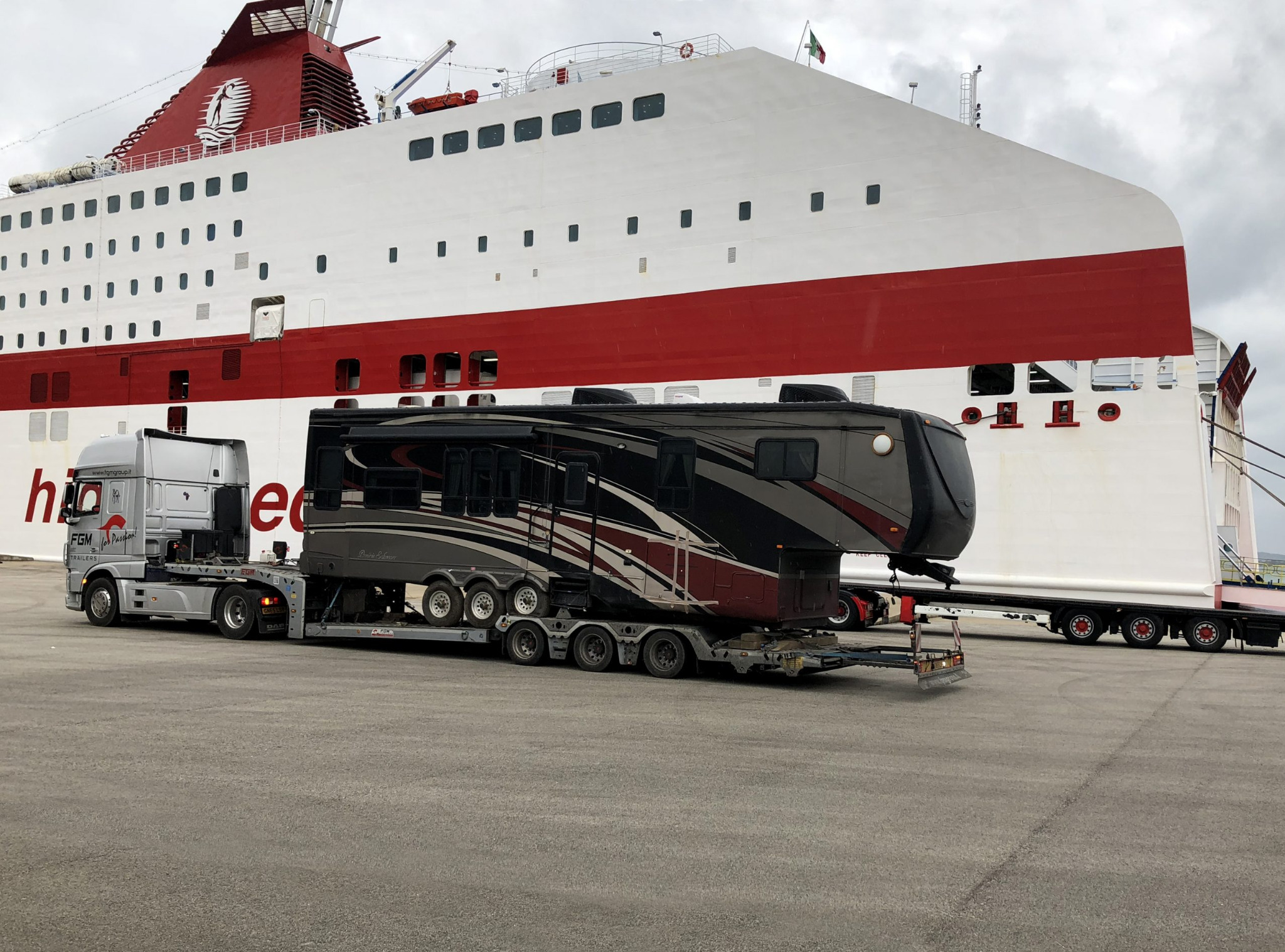 Ciężarówka transportuje dużą przyczepę kempingową na platformie, na tle ogromnego białego promu z czerwonym pasem i włoską flagą.