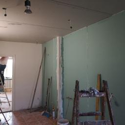 Eugeniusz Pawłowski - Wnętrze w trakcie prac: montaż płyt kartonowo-gipsowych na ścianach i suficie, widoczna drabina, narzędzia malarskie, niedokończone szpachlowanie łączeń płyt.