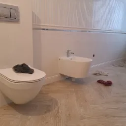 Biała łazienka z bidetem i toaletą w stylu retro, na podłodze jasna drewnopodobna płytka ułożona w jodełkę, na sedesie leży szara ścierka, na ścianach biała boazeria i pionowe panele.