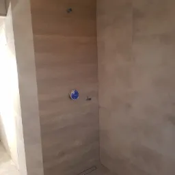 Wykończona ściana prysznicowa z płytek imitujących drewno, z widocznymi przyłączami wodnymi i odpływem liniowym w podłodze, w trakcie prac wykończeniowych łazienki.