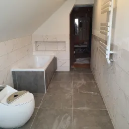 Wykończona łazienka na poddaszu z biało-złotymi płytkami ściennymi, szarą podłogą, wanną w zabudowie i toaletą bez deski, widoczny grzejnik drabinkowy.