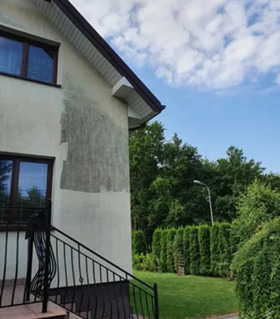 Fragment elewacji domu po częściowym nałożeniu tynku, widoczne schody z metalową balustradą, trawnik i drzewa w tle.