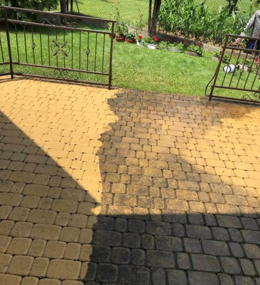 Częściowo wyczyszczona kostka brukowa na tarasie, widoczna różnica w kolorze między czystą a brudną powierzchnią, metalowe ogrodzenie w tle.