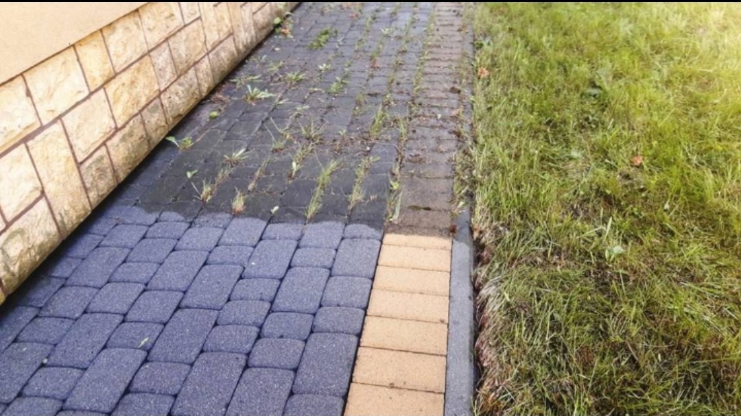 Fragment nawierzchni z kostki brukowej w odcieniach szarości i beżu, częściowo porośniętej chwastami, przylegającej do ściany budynku i trawnika.