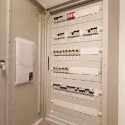 Otwarta szafa rozdzielcza z zabezpieczeniami elektrycznymi, widoczne wyłączniki różnicowoprądowe i nadprądowe, dokumentacja techniczna w przezroczystej okładce po lewej stronie.