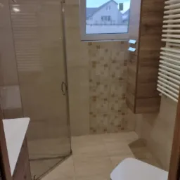 Nowoczesna łazienka z kabiną prysznicową, podwieszaną szafką z drewnianym fornirem, grzejnikiem drabinkowym i widokiem na dom za oknem.