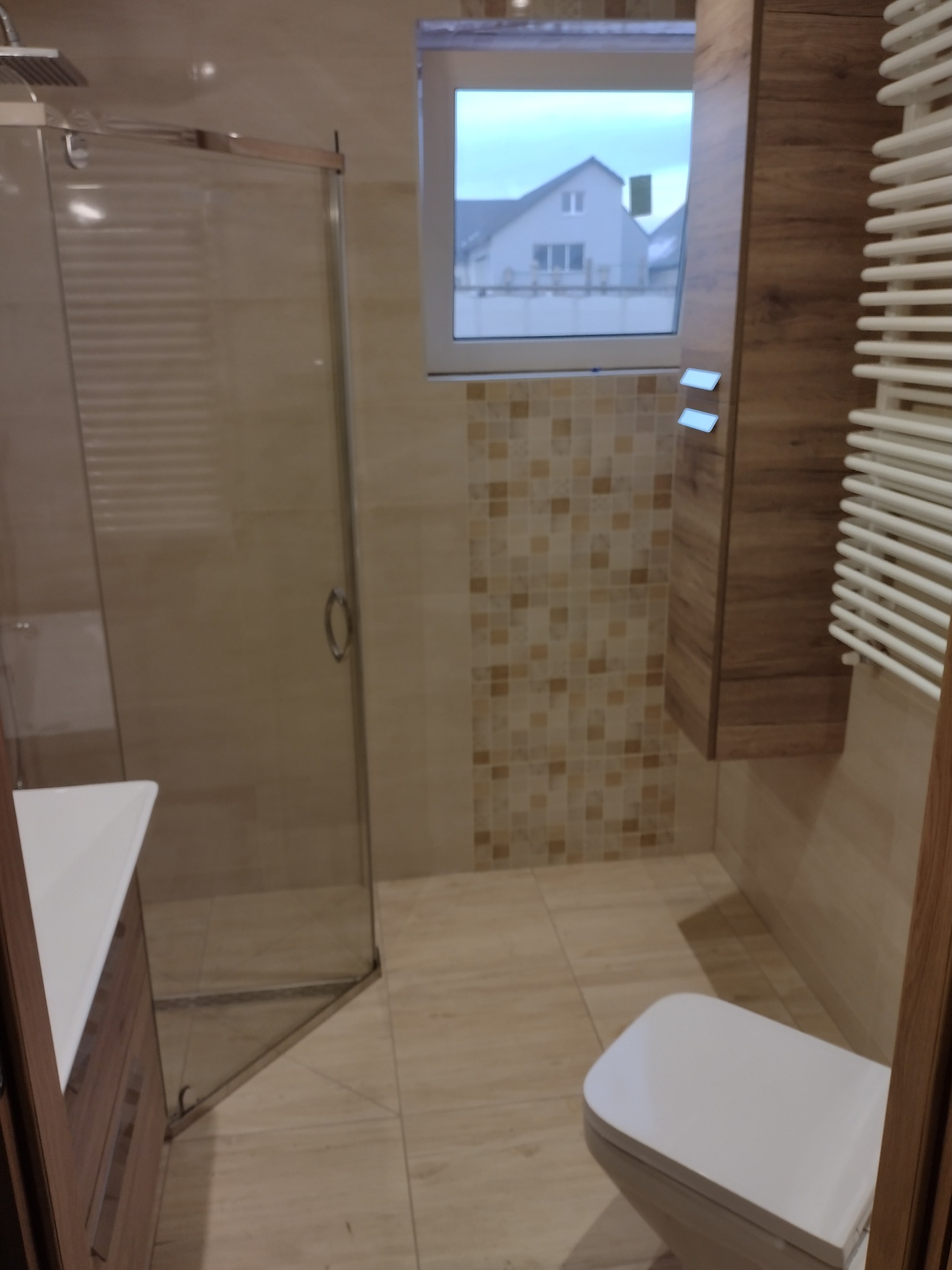 Nowoczesna łazienka z kabiną prysznicową, podwieszaną szafką z drewnianym fornirem, grzejnikiem drabinkowym i widokiem na dom za oknem.