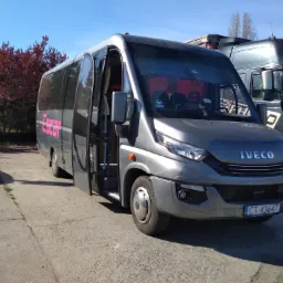 Szary bus Iveco z otwartymi drzwiami na betonowym placu, obok zaparkowana ciężarówka, w tle drzewa i błękitne niebo.