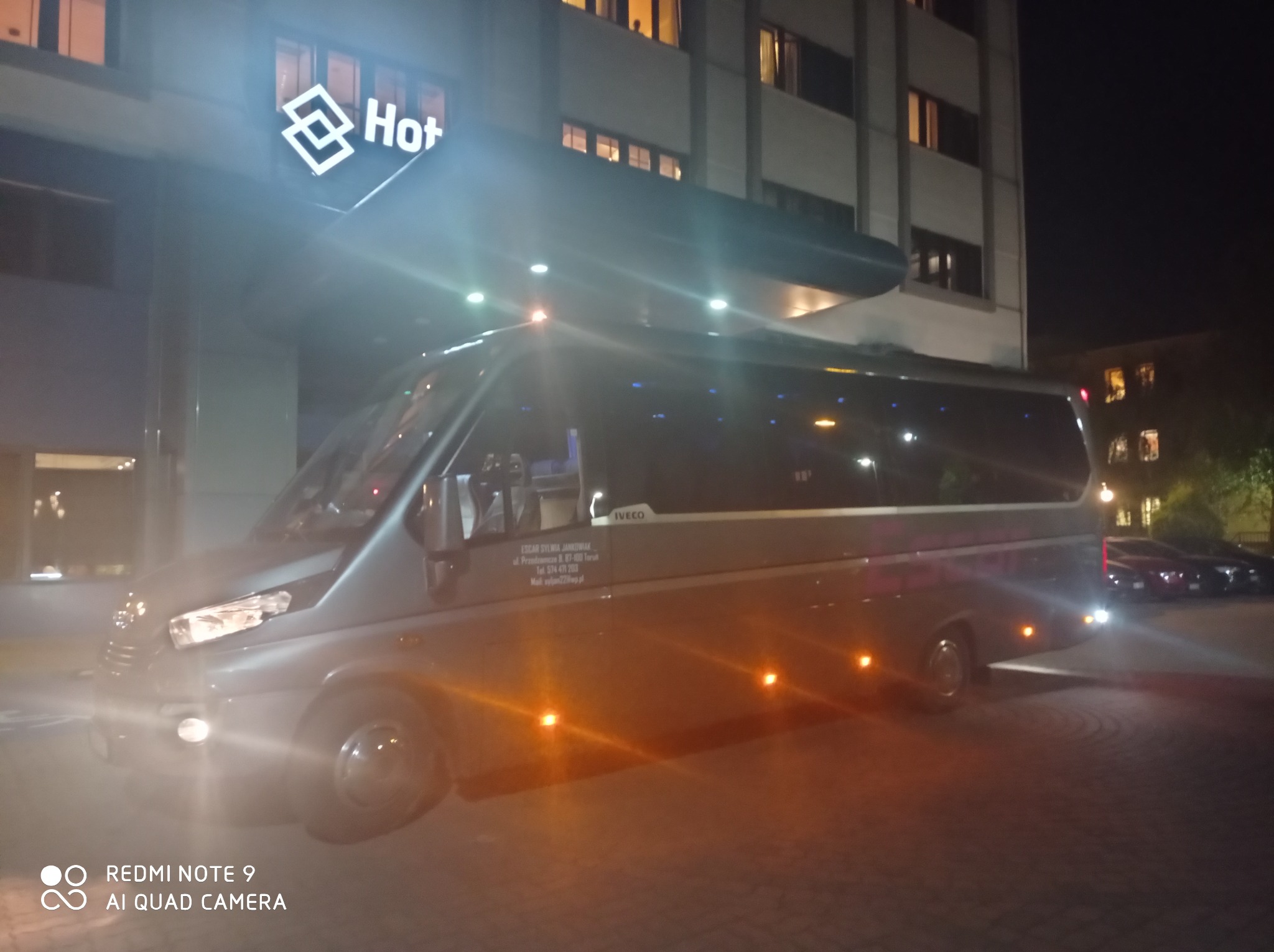 Szary bus marki Iveco zaparkowany nocą przed budynkiem hotelu z podświetlonym logo. Na boku pojazdu widoczne dane kontaktowe firmy transportowej.