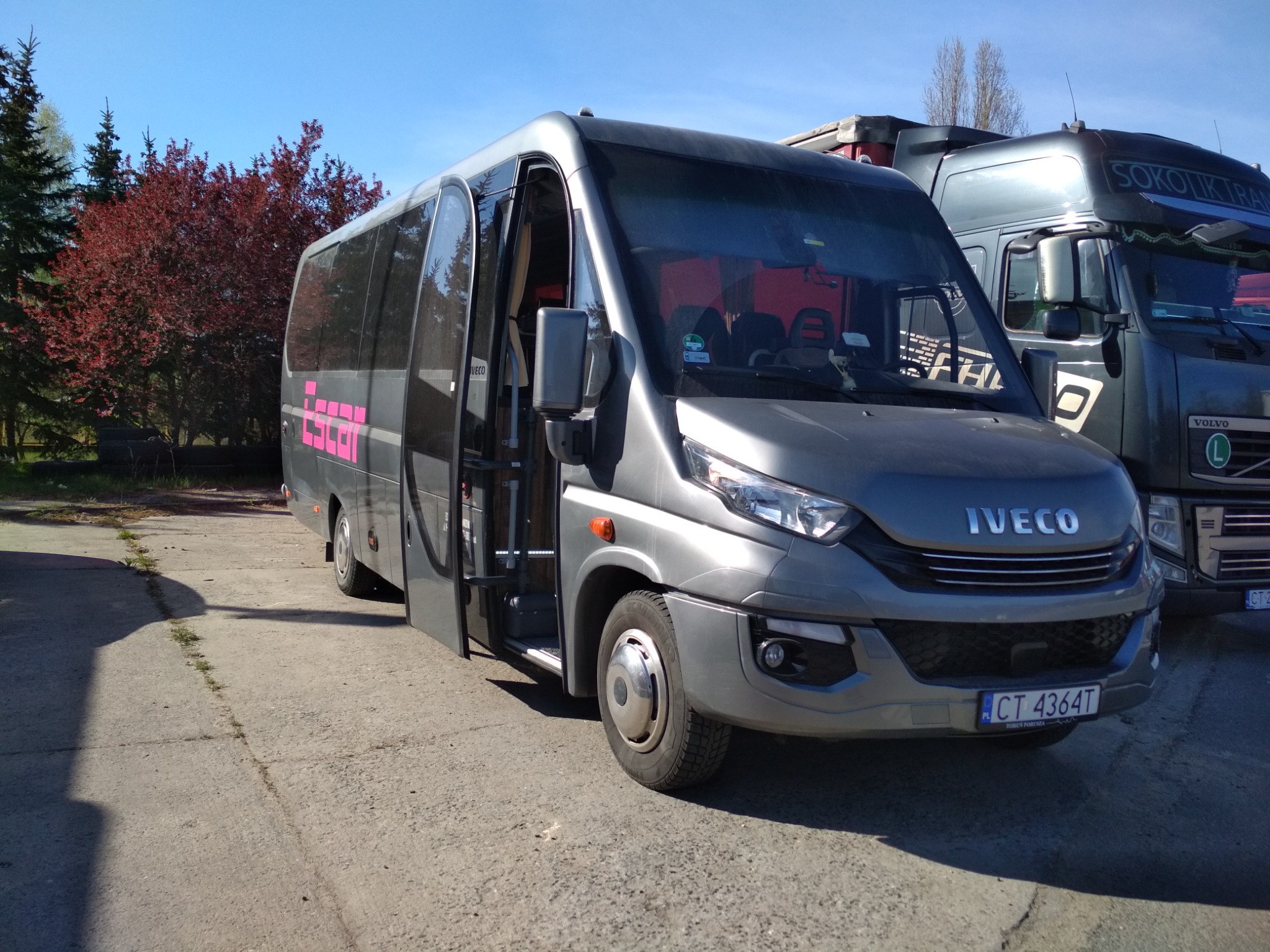 Szary bus Iveco z otwartymi drzwiami na betonowym placu, obok zaparkowana ciężarówka, w tle drzewa i błękitne niebo.