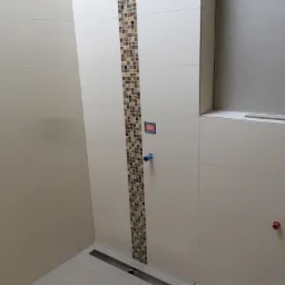 Nowoczesna łazienka w trakcie wykańczania, widoczna jasna glazura ścienna z dekoracyjnym pasem mozaiki, liniowy odpływ podłogowy oraz wystające instalacje wodne przed montażem armatury.