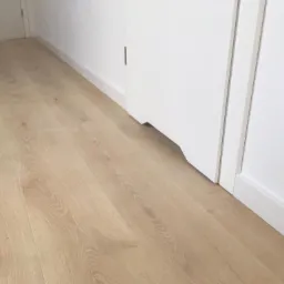 Jasne panele podłogowe ułożone wzdłuż białej ściany z listwą przypodłogową i fragmentem białych drzwi.